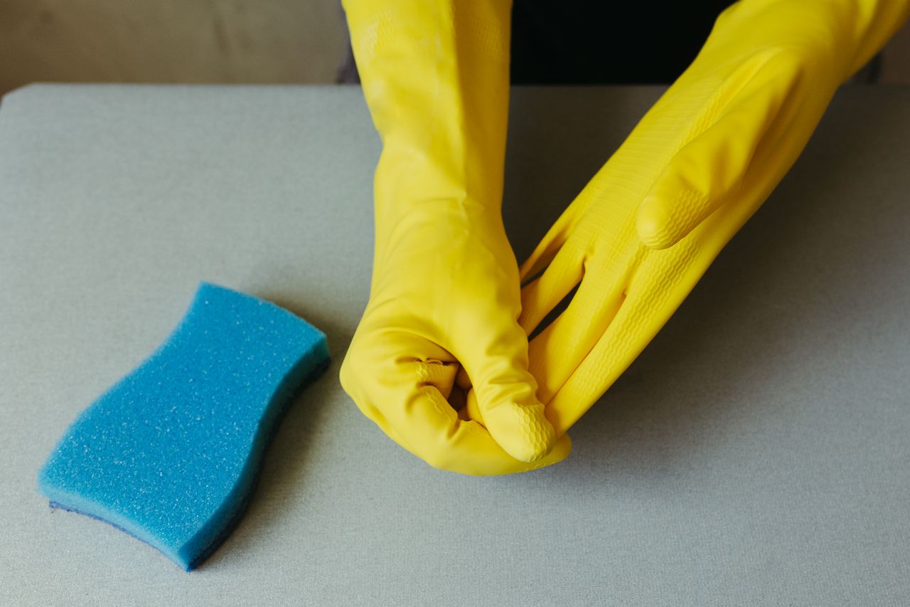 services-02 Close-up of hands in yellow gloves beside a blue sponge on a gray surface.